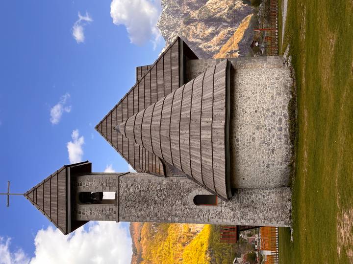 Stone church with wooden shingled roofs set in Alpine valley beneath rocky mountains.