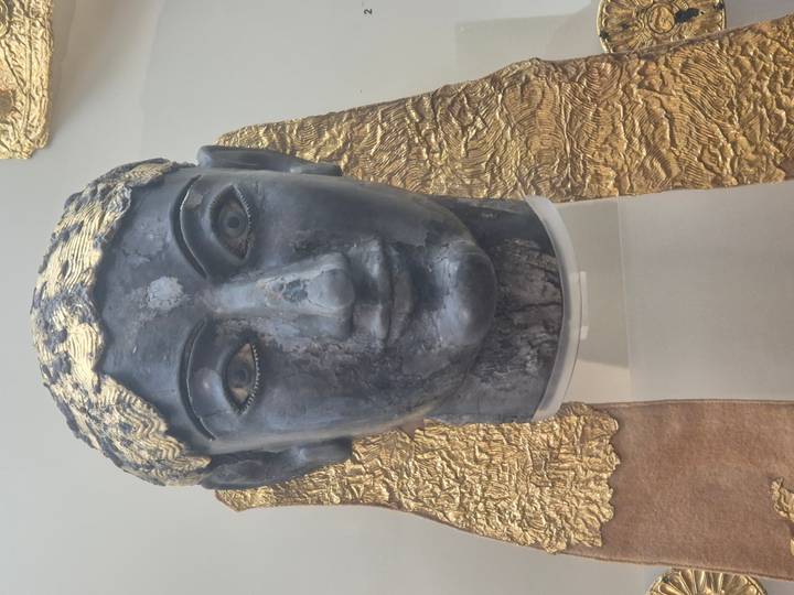 Ancient bronze head adorned with gold leaf displayed in a museum, shown in close detail.