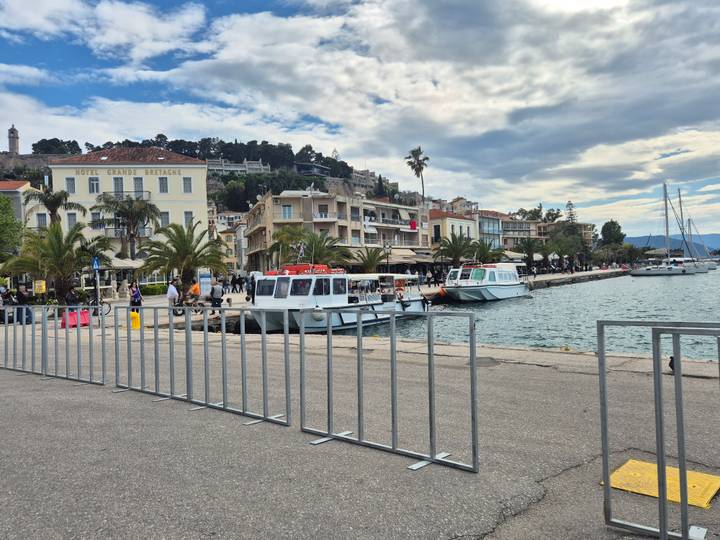 Coastal promenade at Nafplion with moored tour boats, palm trees and historic hilltop fort under mixed clouds.