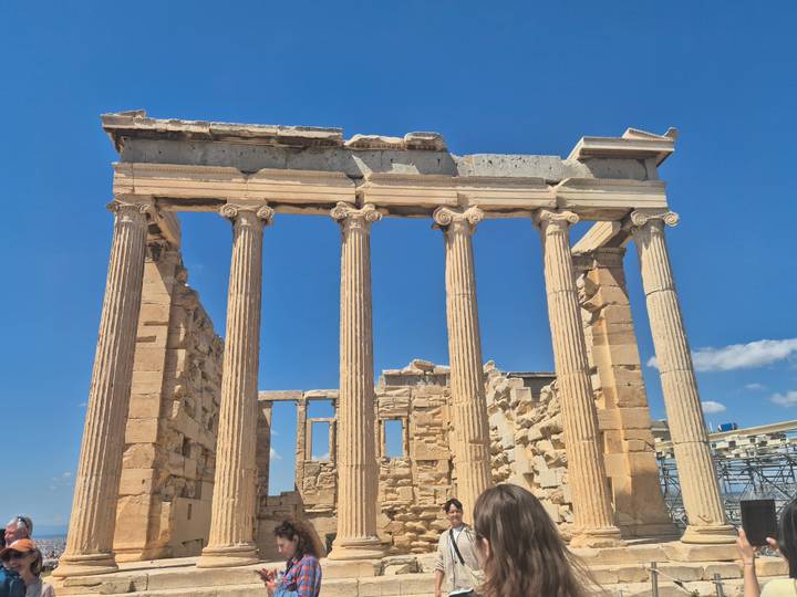 Ruins of the Erechtheion with its tall Ionic columns stand under a cloudless sky on the Acropolis.
