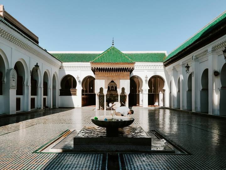 A man bends to drink from a fountain in a serene white-arched courtyard with patterned tile floor.