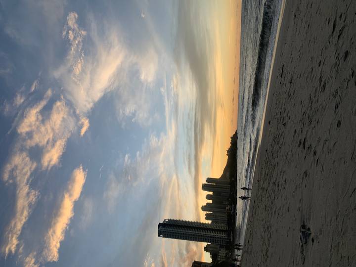 Silhouettes of walkers on a sandy beach with skyscrapers and a pastel sunset sky.