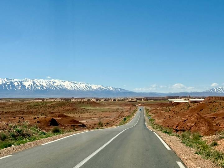 Straight desert highway leads toward distant snow-capped Atlas Mountains beneath clear blue sky.
