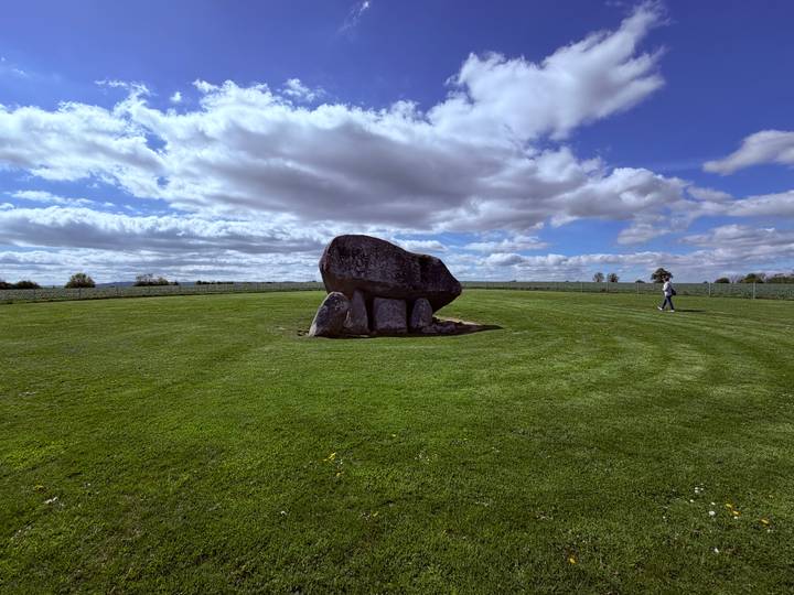 Large ancient dolmen stone structure stands in a vast manicured green field under partly cloudy skies.