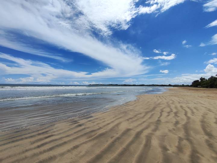 Wide sandy beach with gentle waves and a bright blue sky streaked with dramatic white clouds.