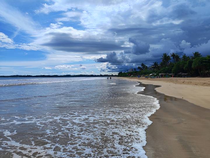 Long stretch of shoreline with gentle surf and dramatic cloud layers over the sea.