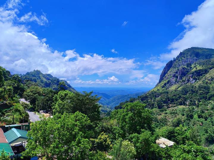 Panoramic mountain valley of lush greenery and distant blue ranges under a brilliant sky.
