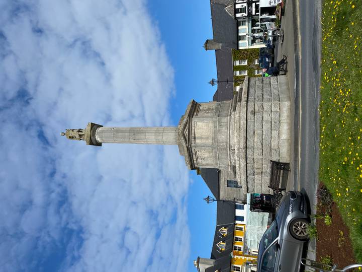 Tall stone column topped with a statue in an Irish town square on a bright day.