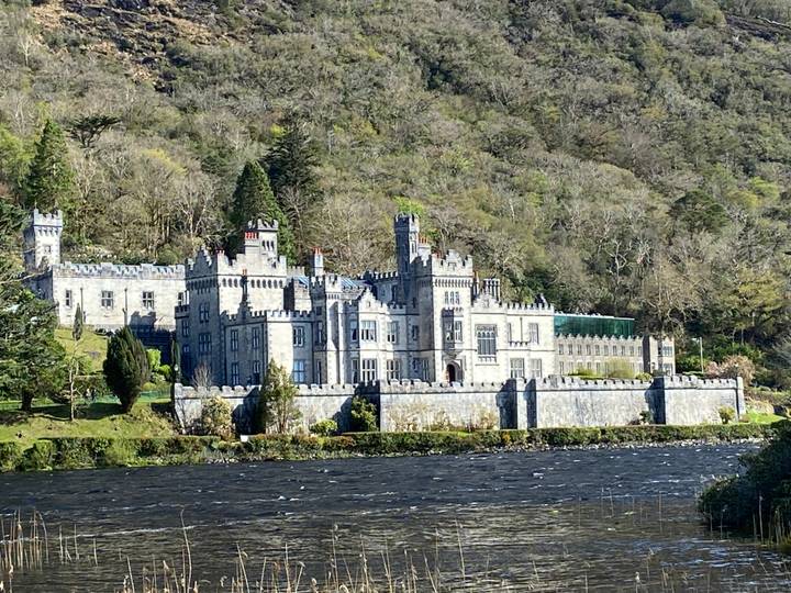 Historic Kylemore Abbey framed by forested hills and a river in the foreground.