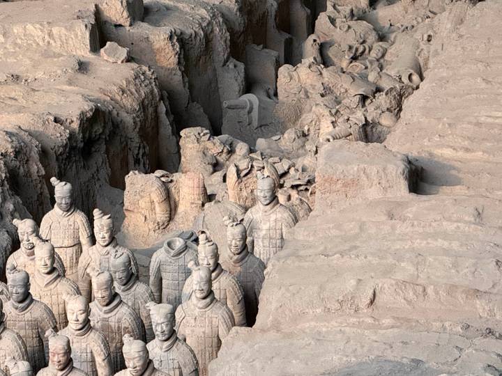 Excavation pit packed with life-sized Terracotta Warriors, some headless and partially unearthed, showcasing intricate armor details in muted clay tones.