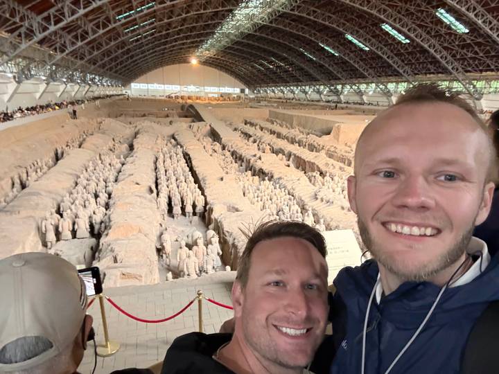 Two smiling travelers take a selfie overlooking the vast Terracotta Army excavation hall filled with warrior statues.