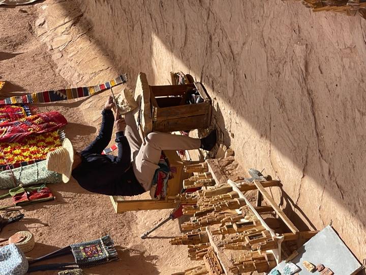 Local artisan in straw hat carving wooden souvenirs in a narrow earthen alleyway.