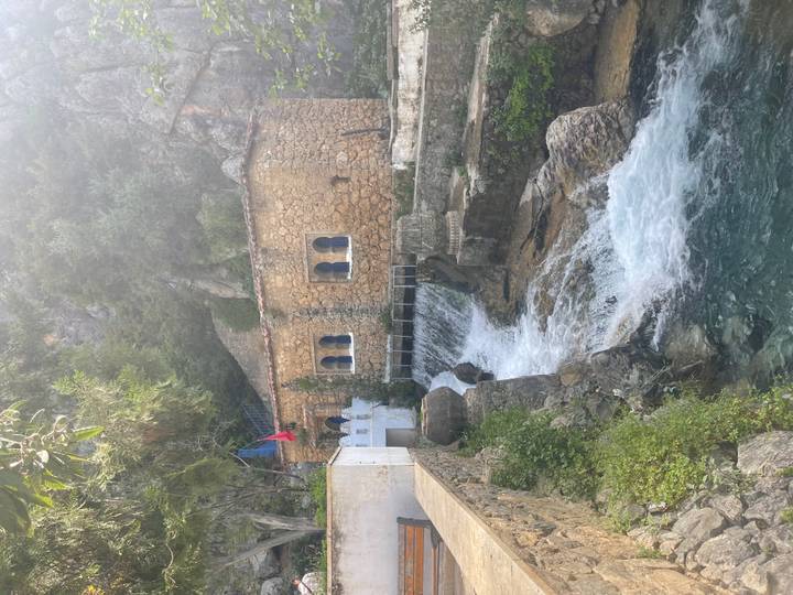 Small stone building beside a cascading stream in a lush, rocky mountain setting with a Moroccan flag.