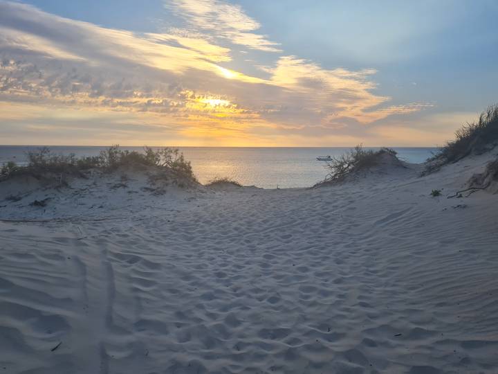 Soft evening light illuminates sand dunes leading to a calm sea with a boat on the horizon.