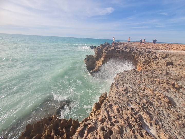 Waves crash into jagged coastal rock formations while a few visitors walk along the cliff top.