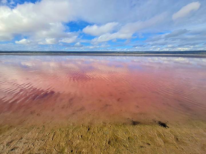 A pink salt lake mirrors clouds and blue sky creating a pastel landscape.