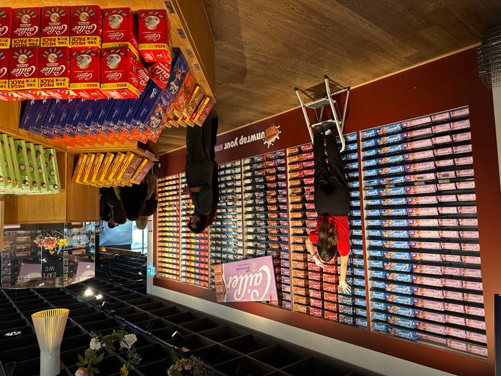 Interior of a Cailler chocolate shop with colourful chocolate bars stacked wall-to-wall and staff stocking shelves.
