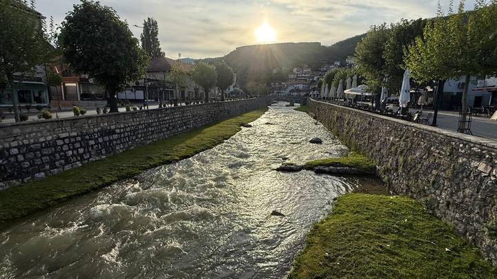 Fast-flowing river flanked by stone embankments and tree-lined walks as the sun sets behind a hilltop town.