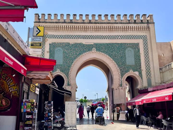 The ornate Bab Bou Jeloud gate in Fes framed by bustling market stalls, pedestrians, and a bright blue sky.