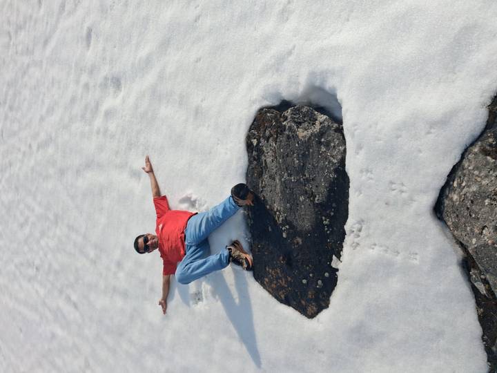 Traveller lies back on snow beside a large dark rock, arms spread wide in playful pose.