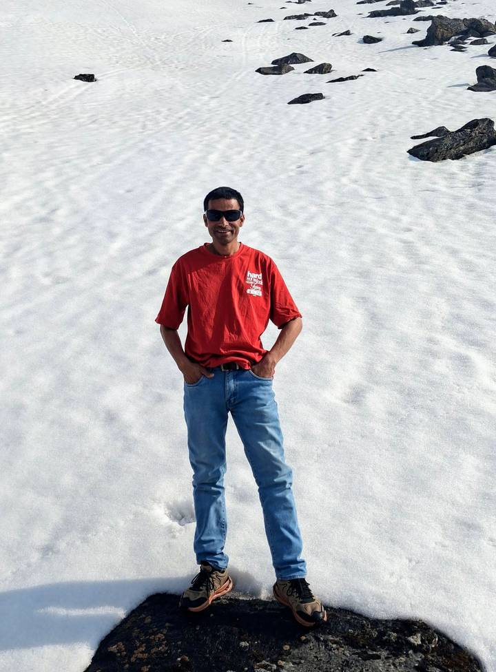 Man poses smiling on a sunlit snowy slope wearing jeans and sunglasses.