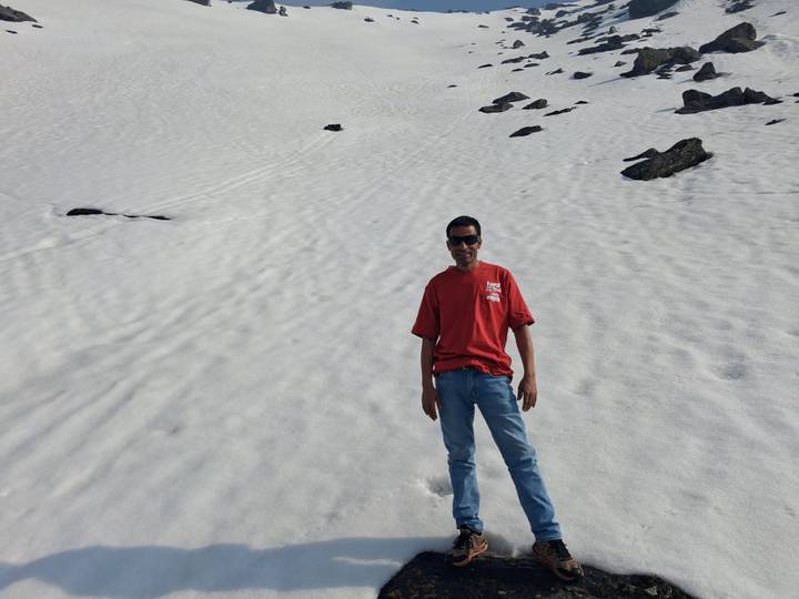 Traveller stands on broad snowfield with scattered dark rocks, distant slope behind.
