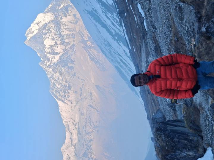 Traveller in bright orange jacket stands before snow-capped Himalayan peaks at dawn.