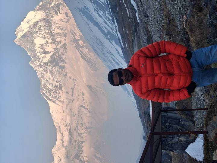 Traveler in red jacket smiling in front of towering icy Himalayan ridge.