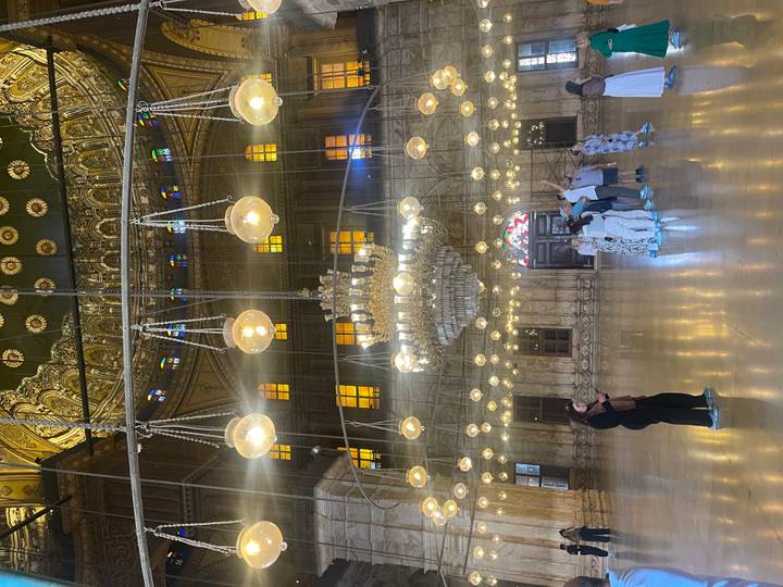 Opulent Ottoman-style mosque interior with hanging globe lamps and marble columns, tourists observing.