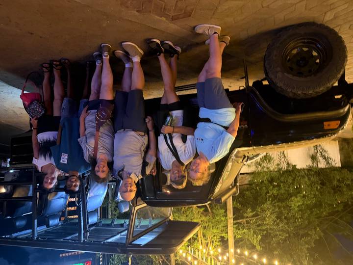 Group of travellers posing beside open safari jeep at night under artificial lights.