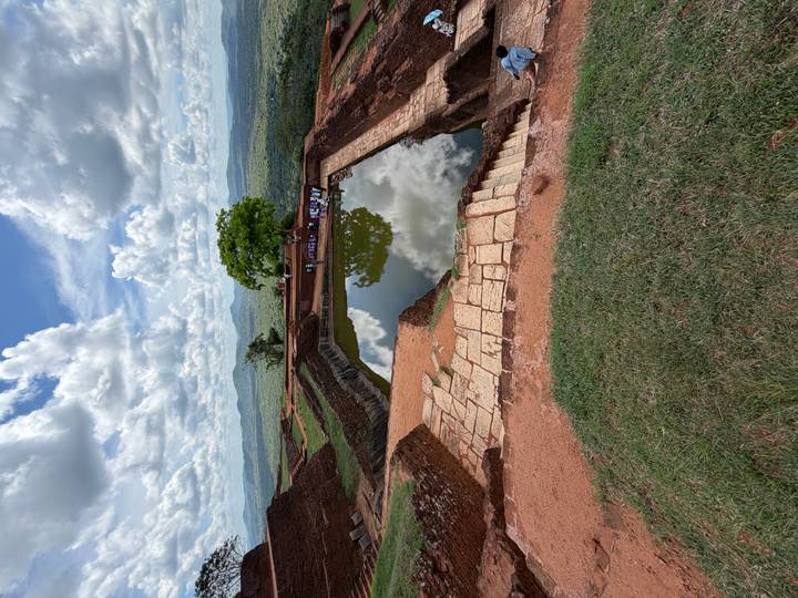 Ancient stone pool atop Sigiriya rock reflecting cloudy sky, surrounded by lush plains.