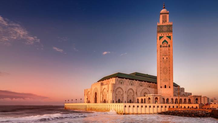 Grand coastal mosque with high minaret bathed in warm sunset light beside gentle ocean waves.