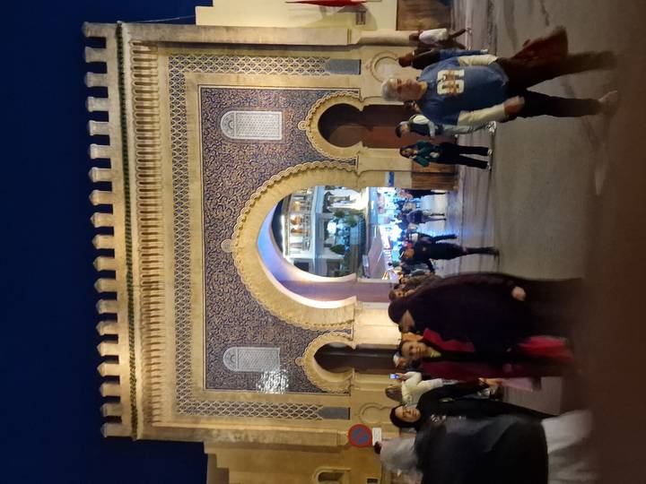 Large ornate mosaic city gate illuminated at dusk with crowds of people walking beneath its arch.