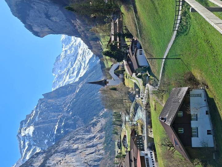 Picturesque Swiss village with church steeple in green valley below towering snow-covered cliffs and peaks.