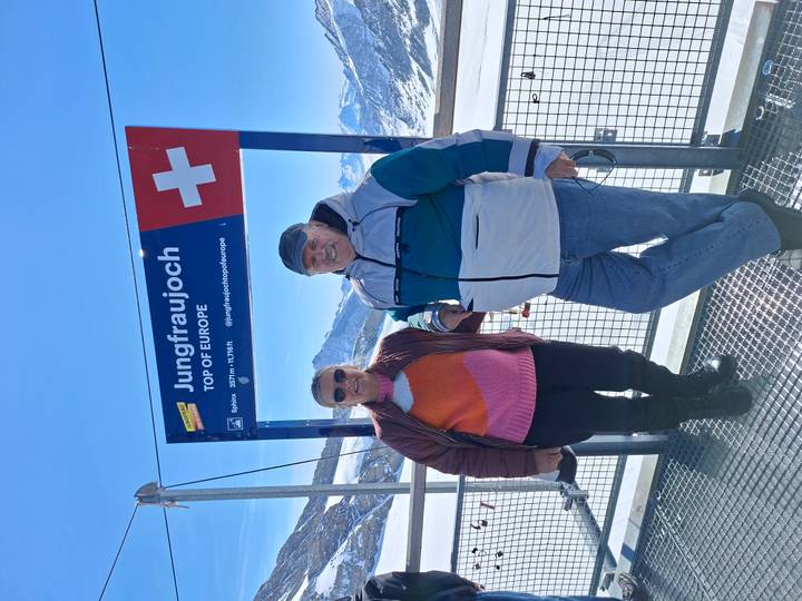 Couple posing at Jungfraujoch observation deck beneath sign 'Top of Europe' with snowy Alps behind.
