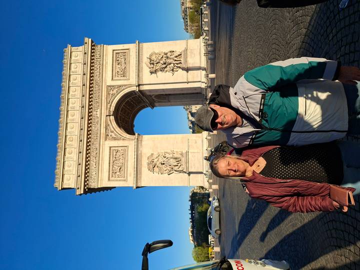 Couple standing in front of the Arc de Triomphe under clear blue sky in late afternoon light.
