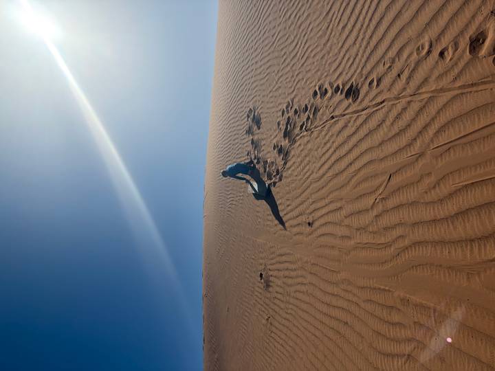 Sandboarder climbing a steep sunlit dune leaving board tracks behind