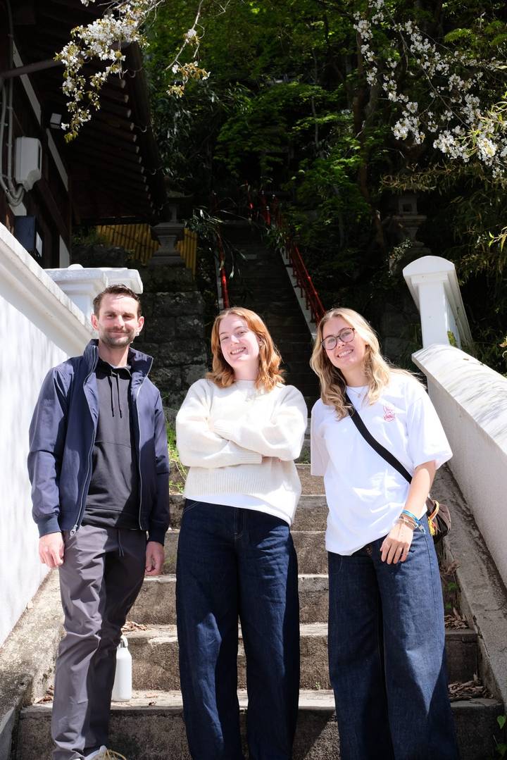 Three travellers smiling on outdoor stone steps with lush greenery and shrine railings behind.