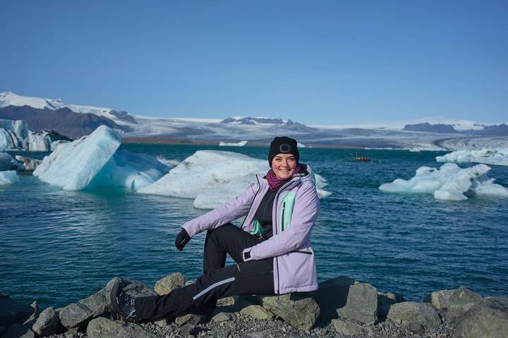 Smiling traveler in winter gear seated on rocks by an ice-filled glacial lagoon under blue sky