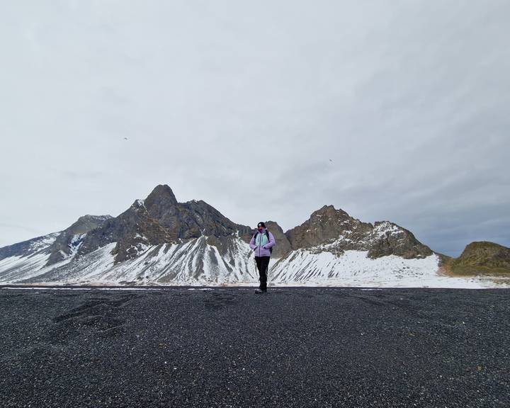 Solo hiker on a black sand plain with snow-striped jagged peaks rising behind