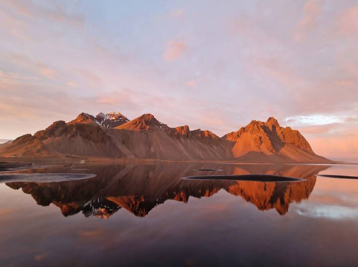 Dramatic orange sunrise light reflecting jagged mountains in still lagoon water at Vestrahorn