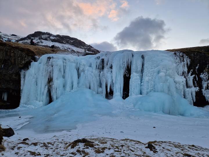 Frozen waterfall with thick blue ice columns beneath a snowy hillside at dusk