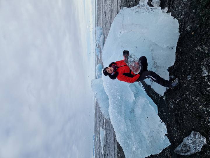 Traveler in red jacket kneeling on clear blue ice shards on a volcanic black beach