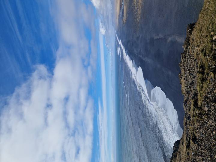 Long aerial view of black sand coastline with white surf and vast blue ocean under scattered clouds