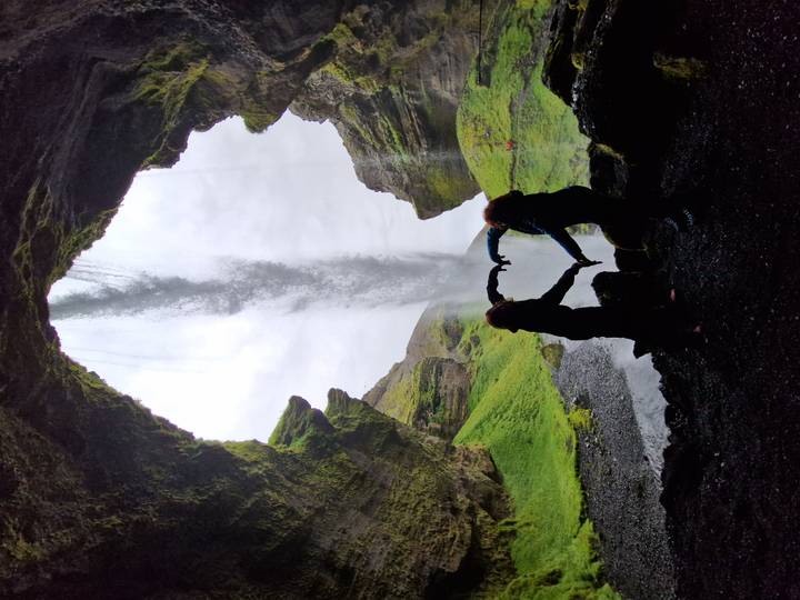 Two silhouetted travelers forming a heart shape inside a cave behind a waterfall curtain