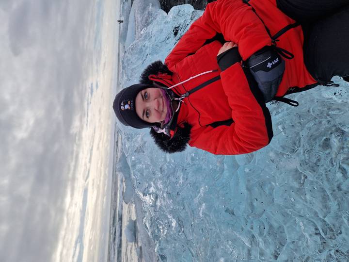 Young woman in a red jacket leans against a vivid blue ice block at a glacial beach under a cloudy sky.