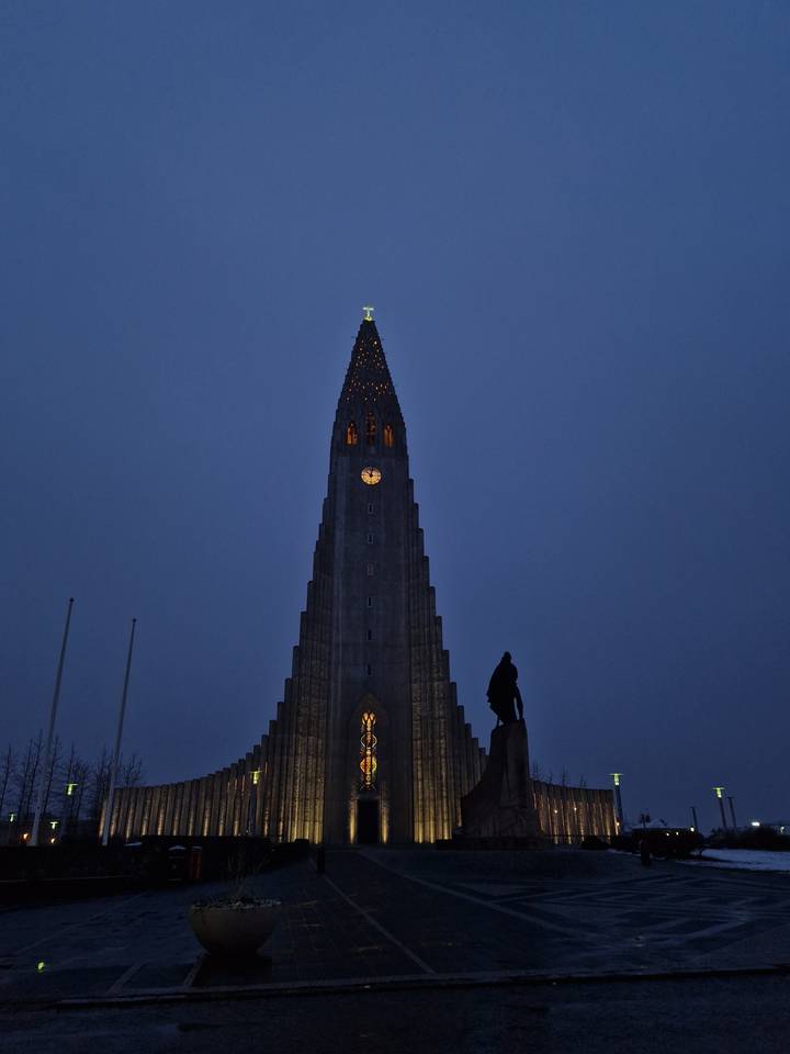 Hallgrimskirkja church tower lit up against a deep blue evening sky with a small statue silhouette in front.