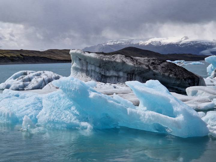 Jagged blue and black icebergs float in turquoise water beneath cloudy skies and distant snowy peaks.
