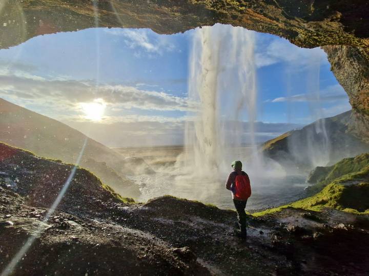 Traveler in red jacket stands inside a misty cave behind a waterfall as sunlight streams through.