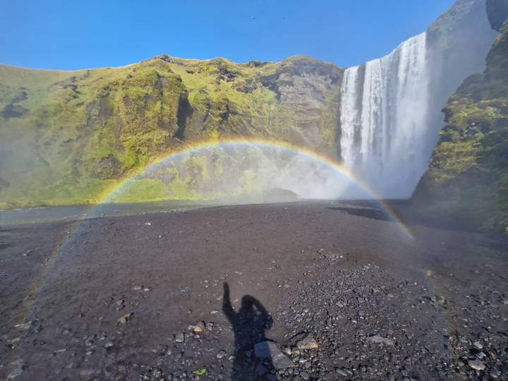 Rainbow arcs in mist before a tall waterfall plunging from mossy cliffs on a bright day.